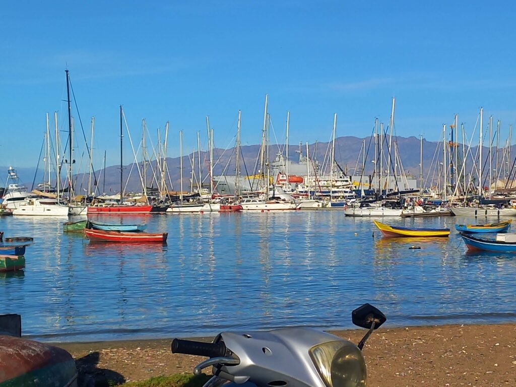 Yachthafen vor Bergkulisse mit vielen Masten; bunte Ruderboote im ruhigen Wasser, Roller steht am Kiesufer