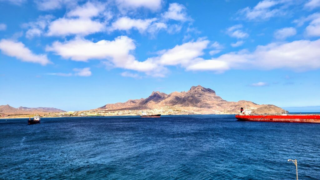 Blaues Meer vor karger Insel mit Bergen; roter Frachter rechts, zwei kleinere Schiffe links unter lockerem Wolkenhimmel.