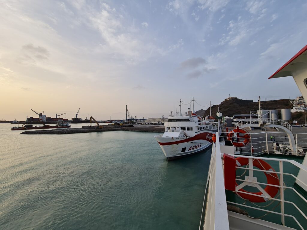Einfahrt zum Hafen bei Sonnenuntergang: weiß-rotes Armas-Fährschiff angelegt, ruhiges türkisfarbenes Wasser, Kräne im Hintergrund