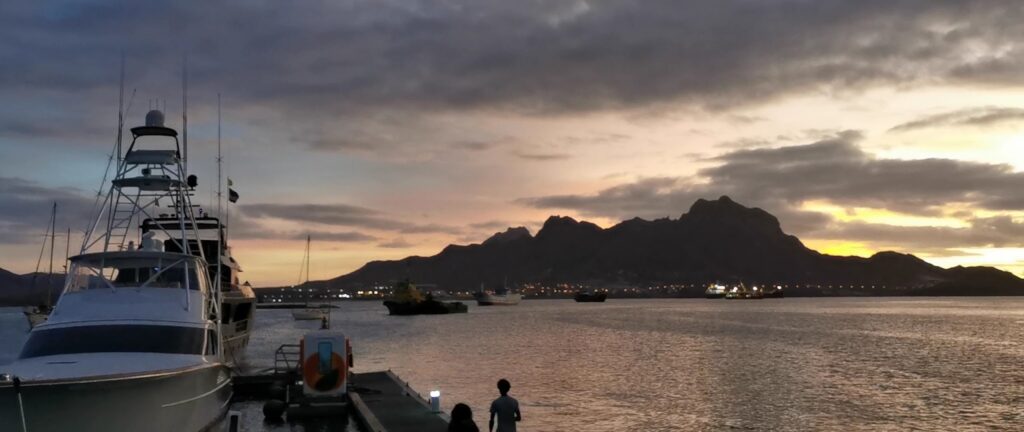 Yacht am Pier, zwei Personen schauen aufs Wasser; dahinter beleuchteter Hafen, dunkle Bergsilhouette im Sonnenuntergang.