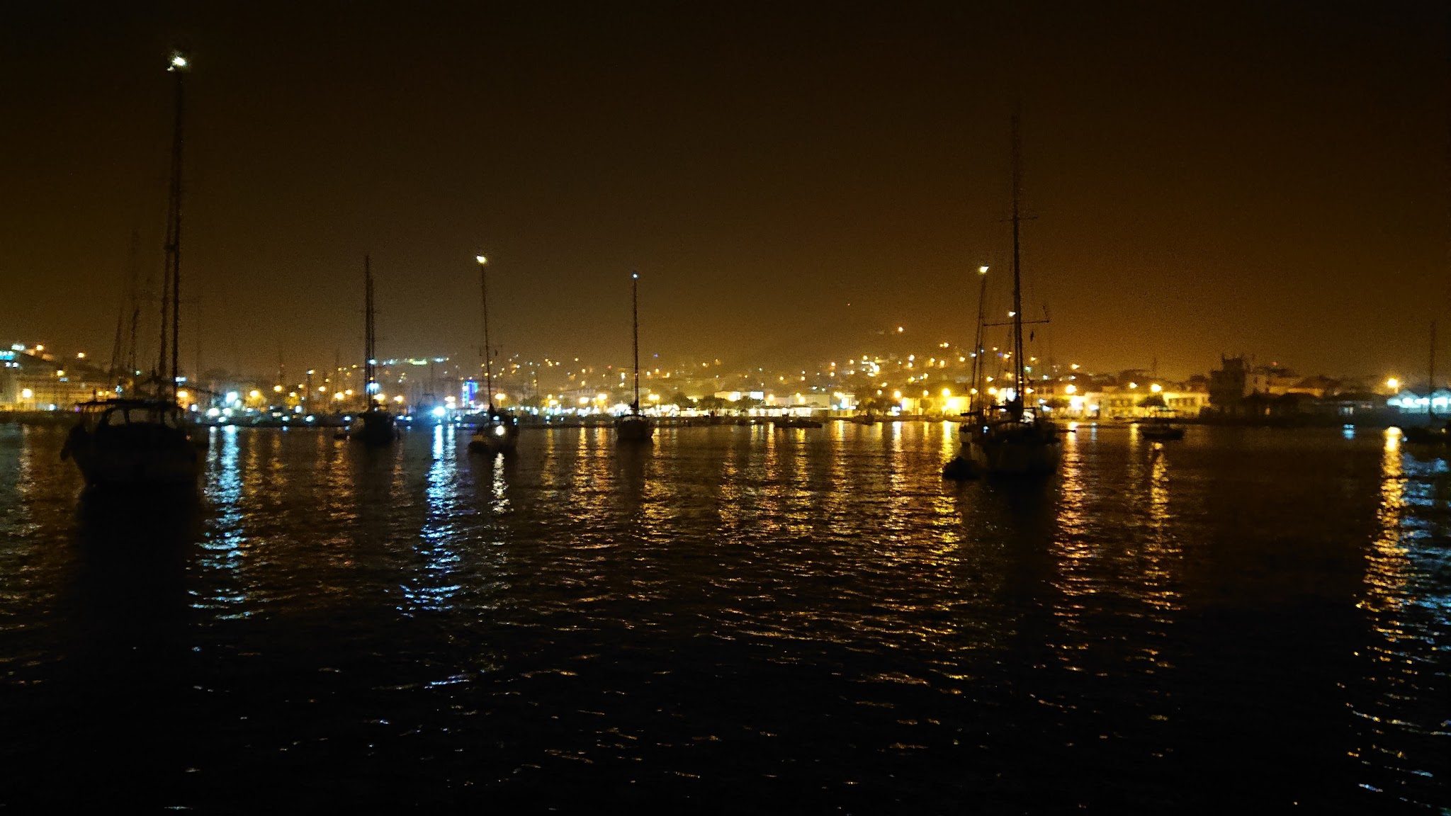 Silhouettierte Segelboote liegen nachts im Hafen, orange Stadtlichter spiegeln sich auf dunklem Wasser.