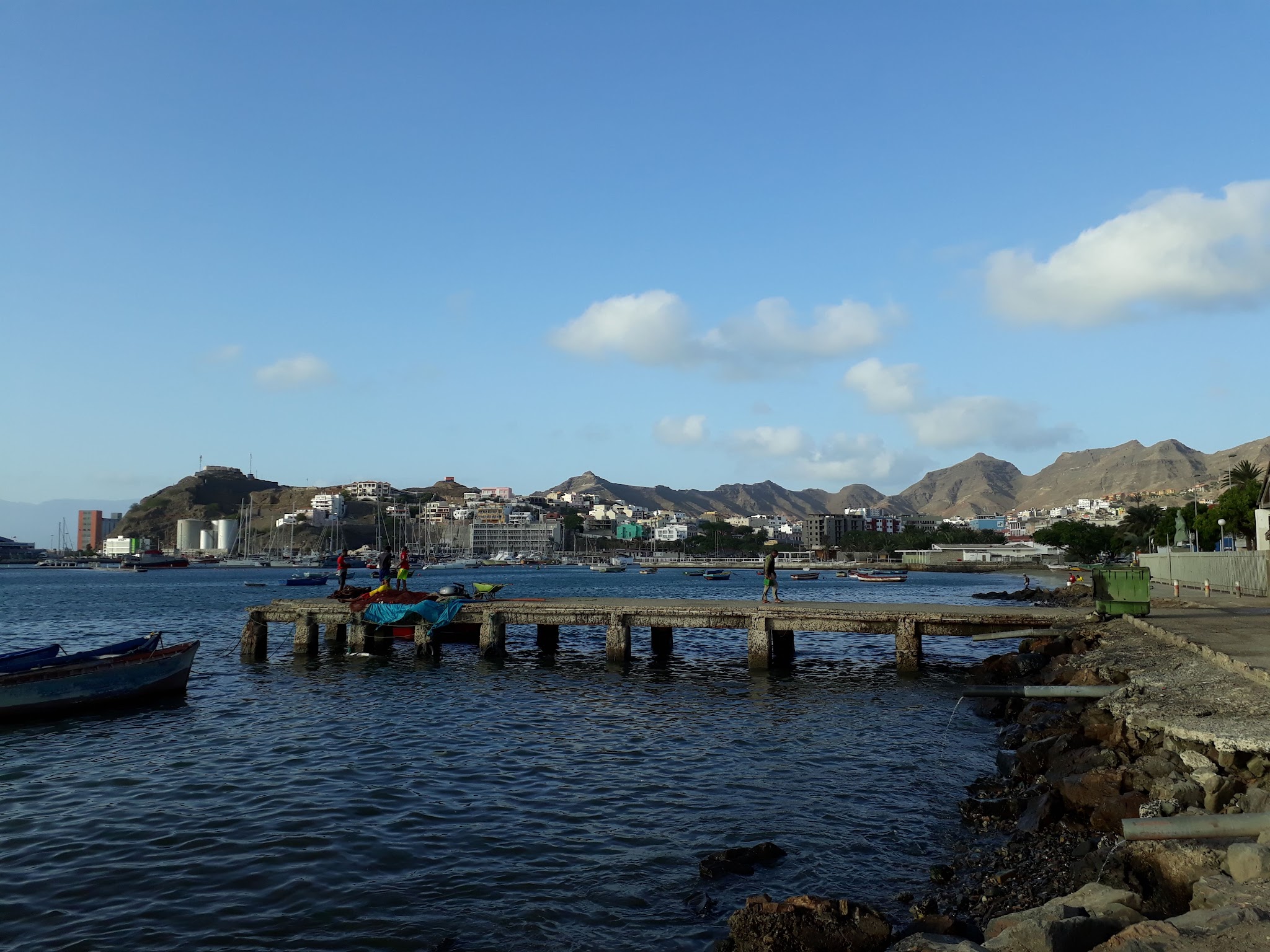 Kleiner Pier mit Fischern und bunten Booten vor Stadt am Hafen; dahinter karge Berge unter blauem Himmel.