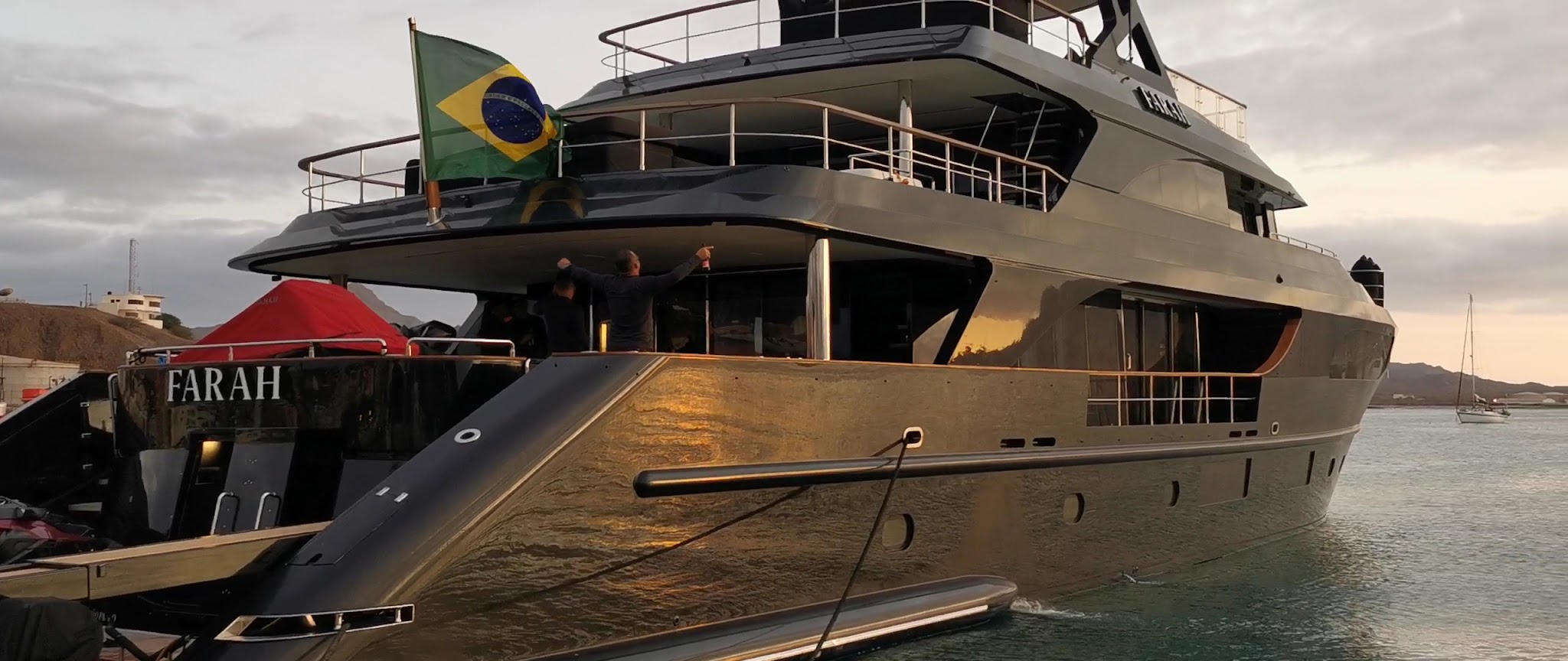 Luxusmotoryacht FARAH mit wehender brasilianischer Flagge im Hafen, zwei Personen am Heck bei Sonnenuntergang.
