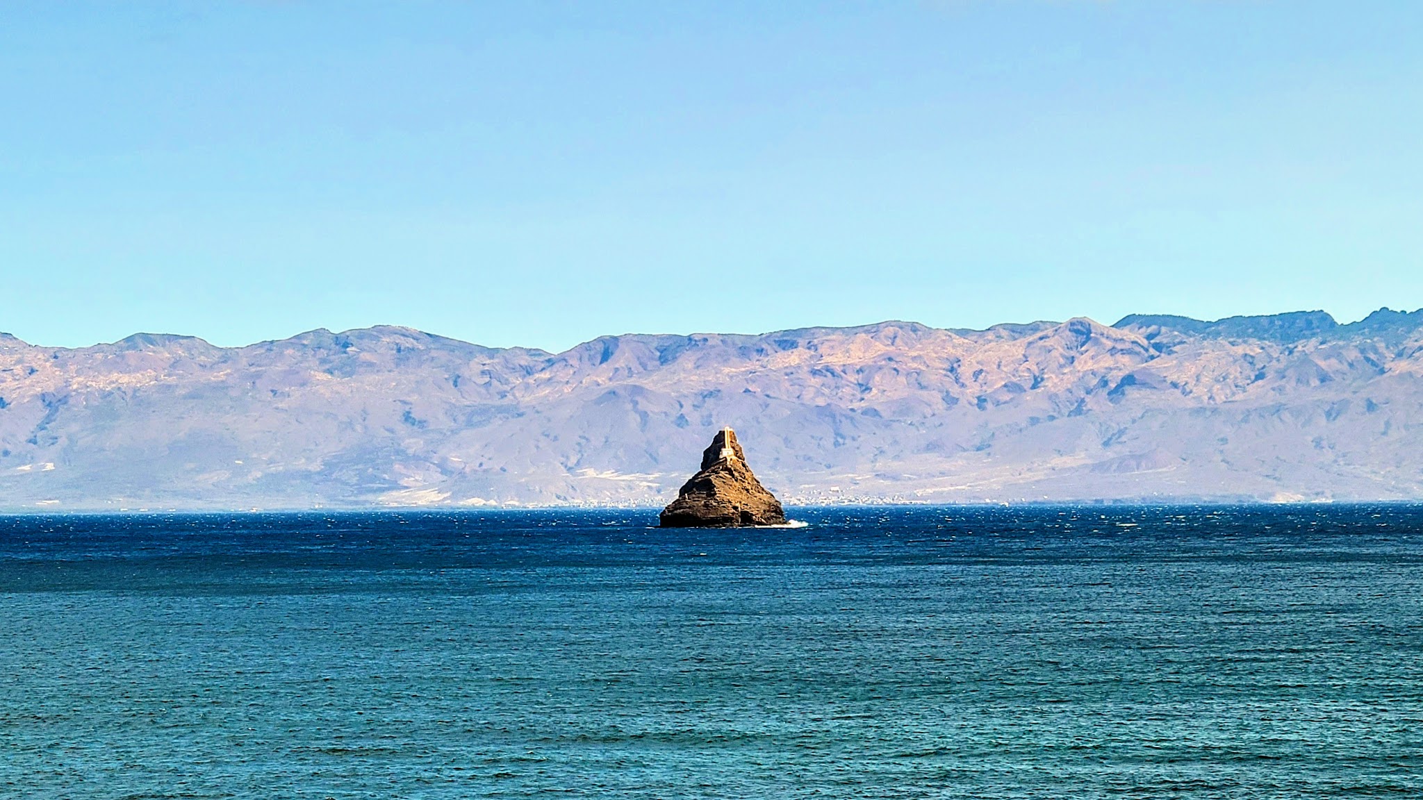 Einzelner spitzer Felsen erhebt sich aus türkisblauem Meer, dahinter sandbraune Bergkette unter wolkenlosem Himmel.