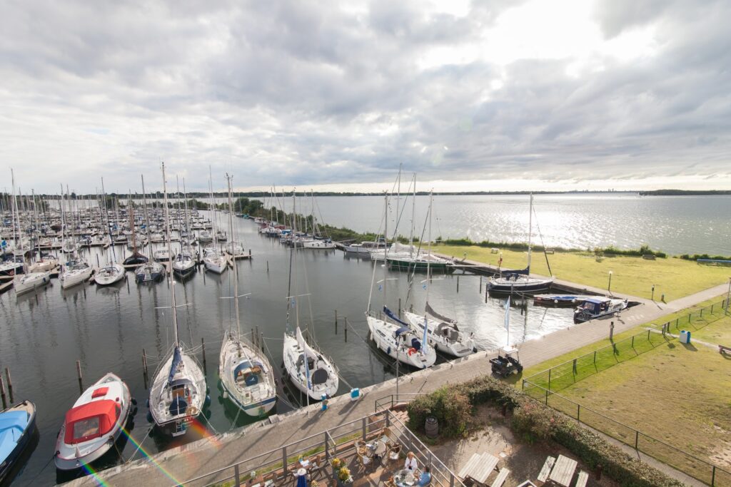 Segelboote dicht an Stegen, einige Yachten im Vordergrund; ruhiger See glitzert unter bewölktem Himmel.