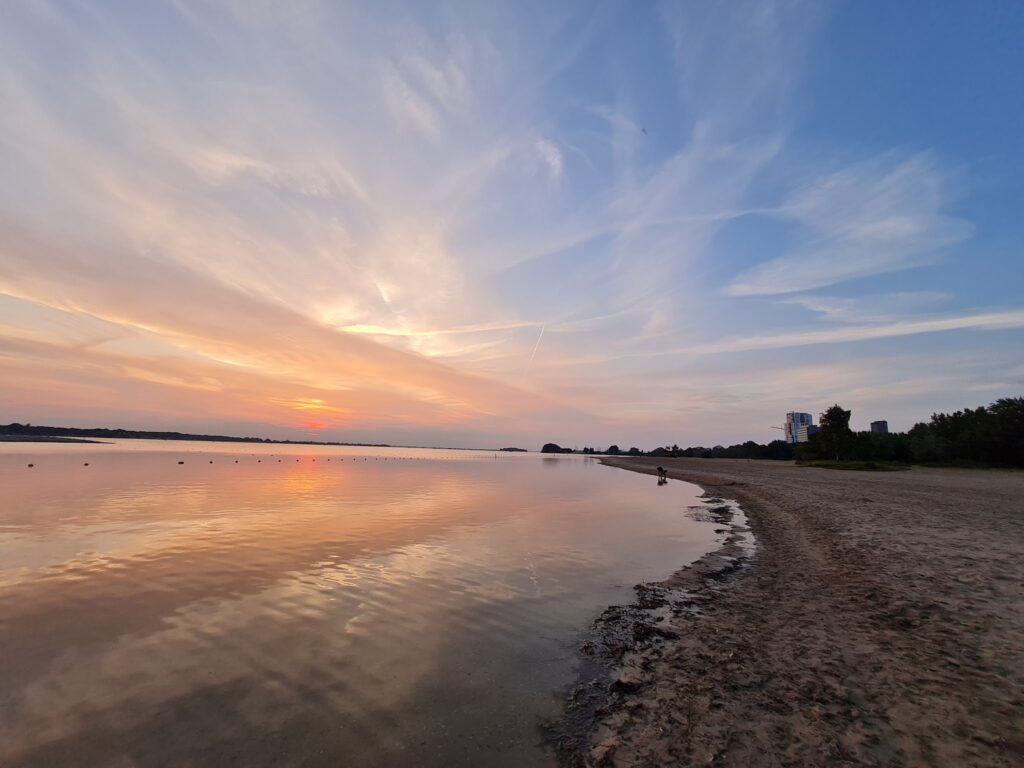 Ruhiger See bei Sonnenuntergang, orange-rosa Himmel spiegelt sich im Wasser; sandiges Ufer, ferne Bäume und Hochhaus