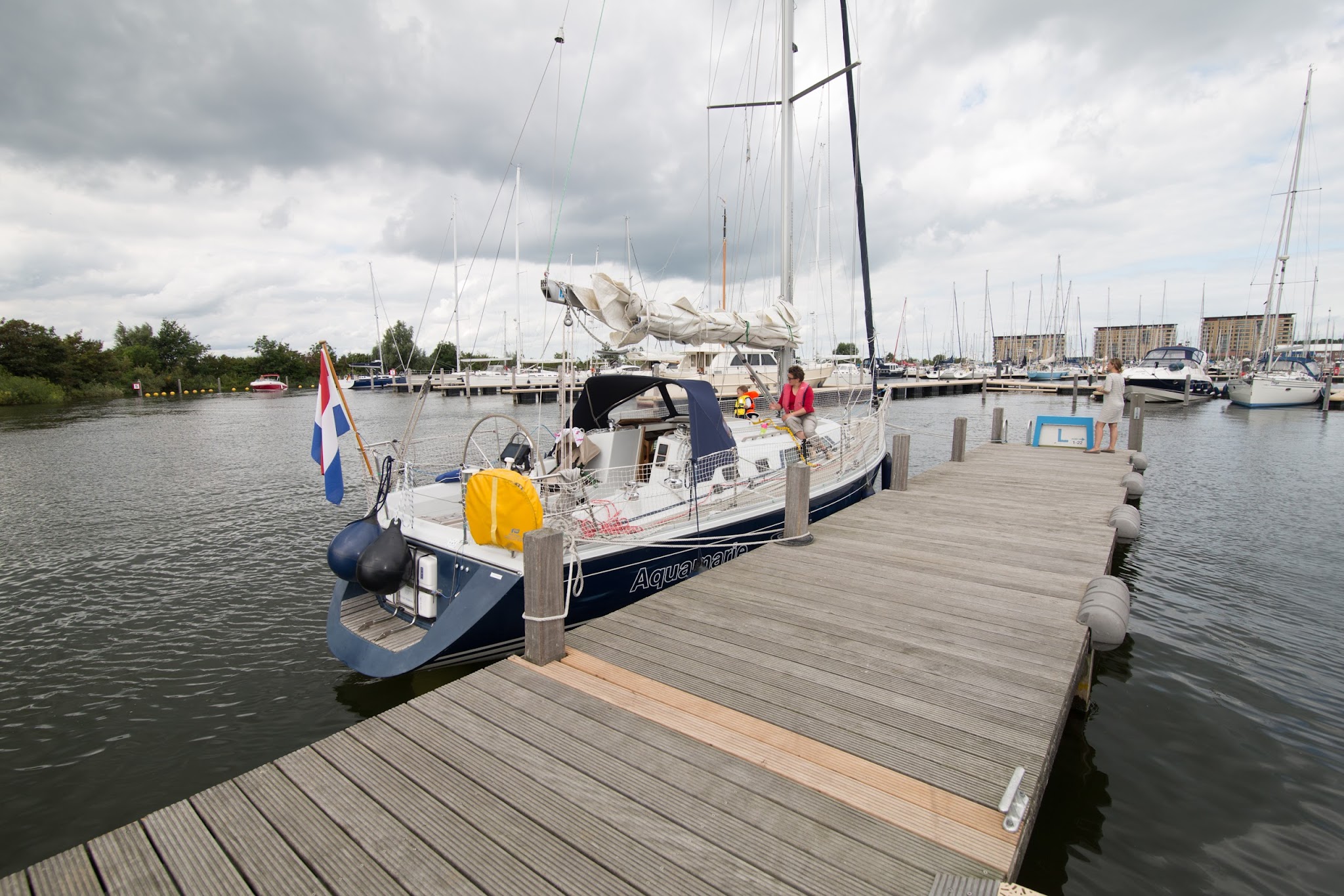 Segelboot mit niederländischer Flagge liegt am Holzsteg; Person in pink sitzt an Deck, Yachthafen im Hintergrund.