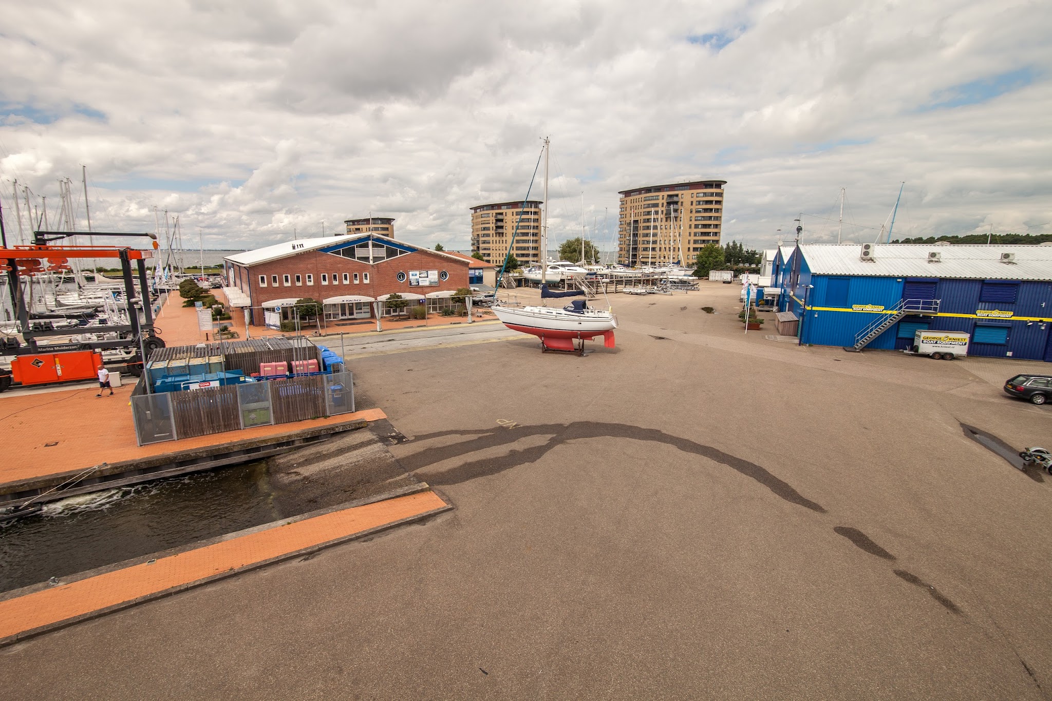 Segelboot steht auf Böcken im Yachthafen zwischen Backsteingebäude, blauen Hallen und Hochhäusern unter Wolkenhimmel