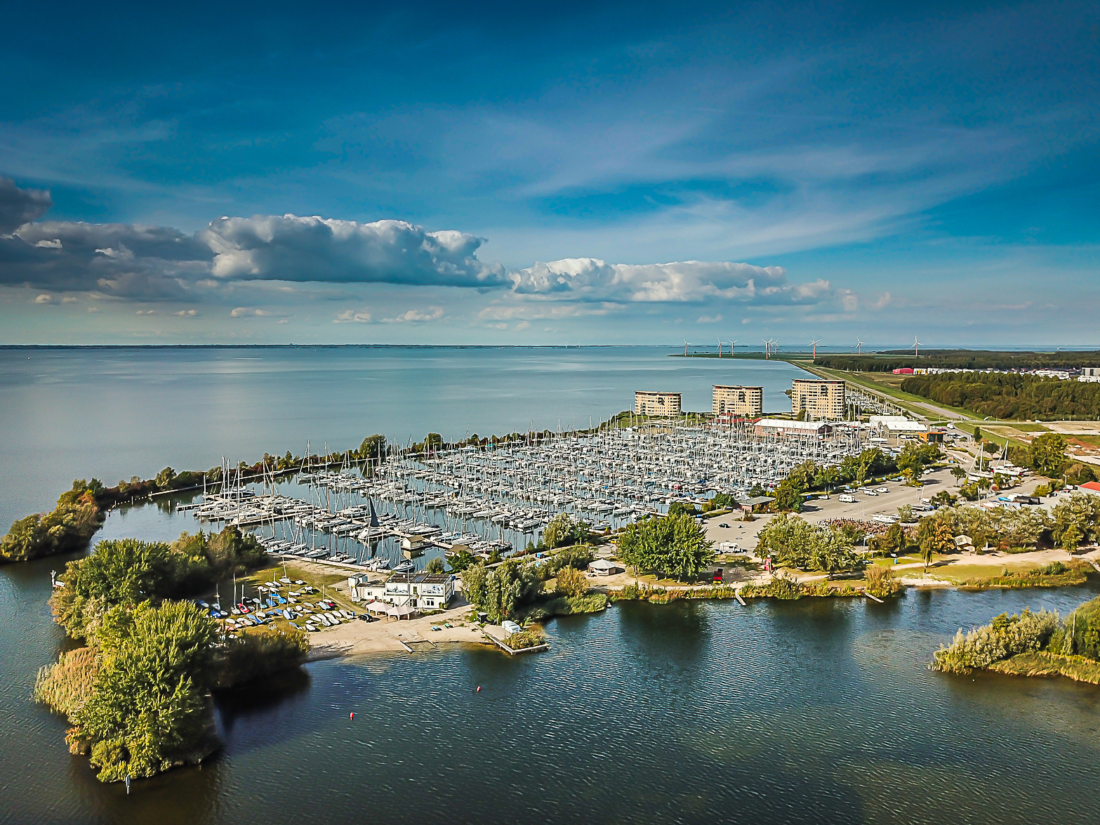 Luftaufnahme eines großen Jachthafens voller Segelboote an ruhigem See; im Hintergrund Deich, Windräder und Wolkenhimmel