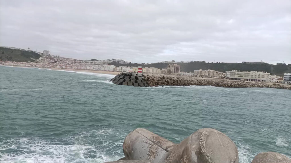 Grauer Himmel über einer Küstenstadt; Wellen schlagen gegen einen Wellenbrecher mit rot-weiß gestreiftem Leuchtturm.