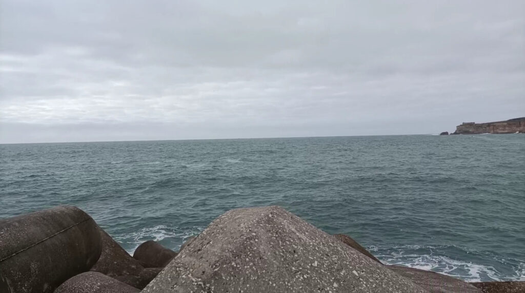 Grau bewölkter Himmel über welligem Meer; vorn Betonwellenbrecher, rechts fern felsige Klippe