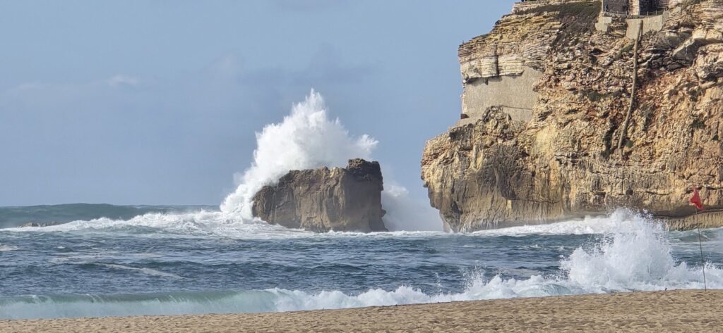 Hohe Welle schlägt gegen Felsen vor steiler Klippe; rote Warnflagge am Strand im Vordergrund.