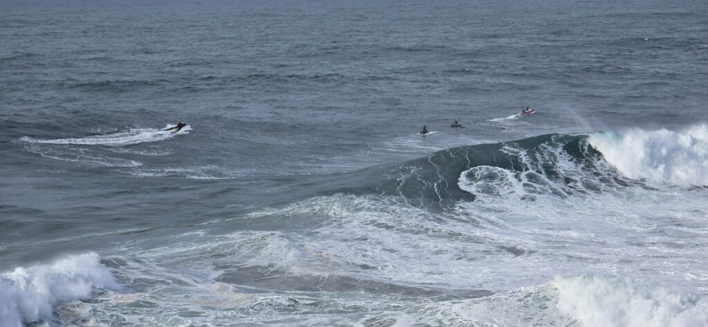 Vier Jetskis in unruhiger See vor einer brechenden Riesenwelle.