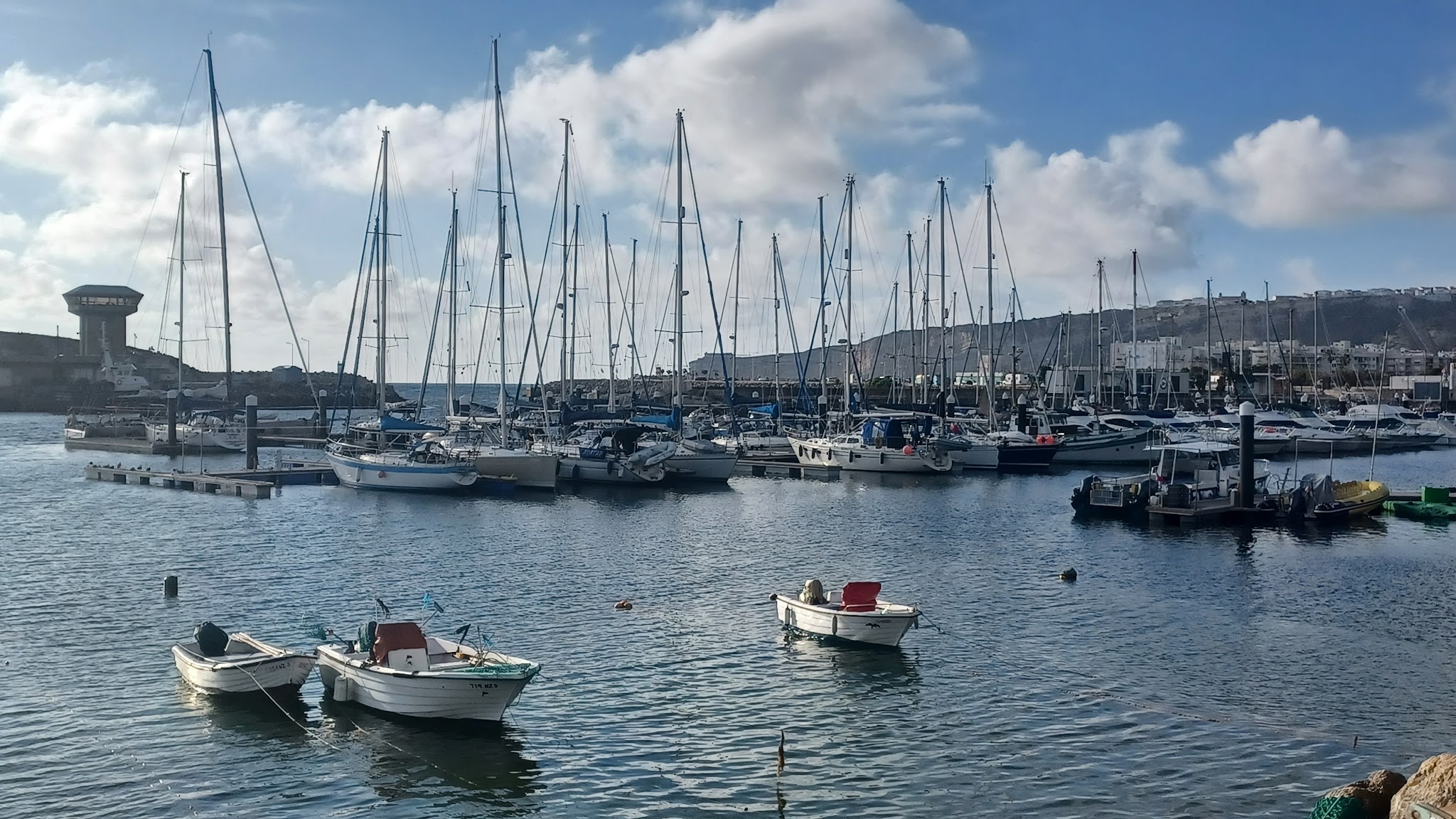 Yachthafen bei Tag: dicht aneinandergereihte Segelboote, drei kleine Boote im Vordergrund, blauer Himmel mit Wolken