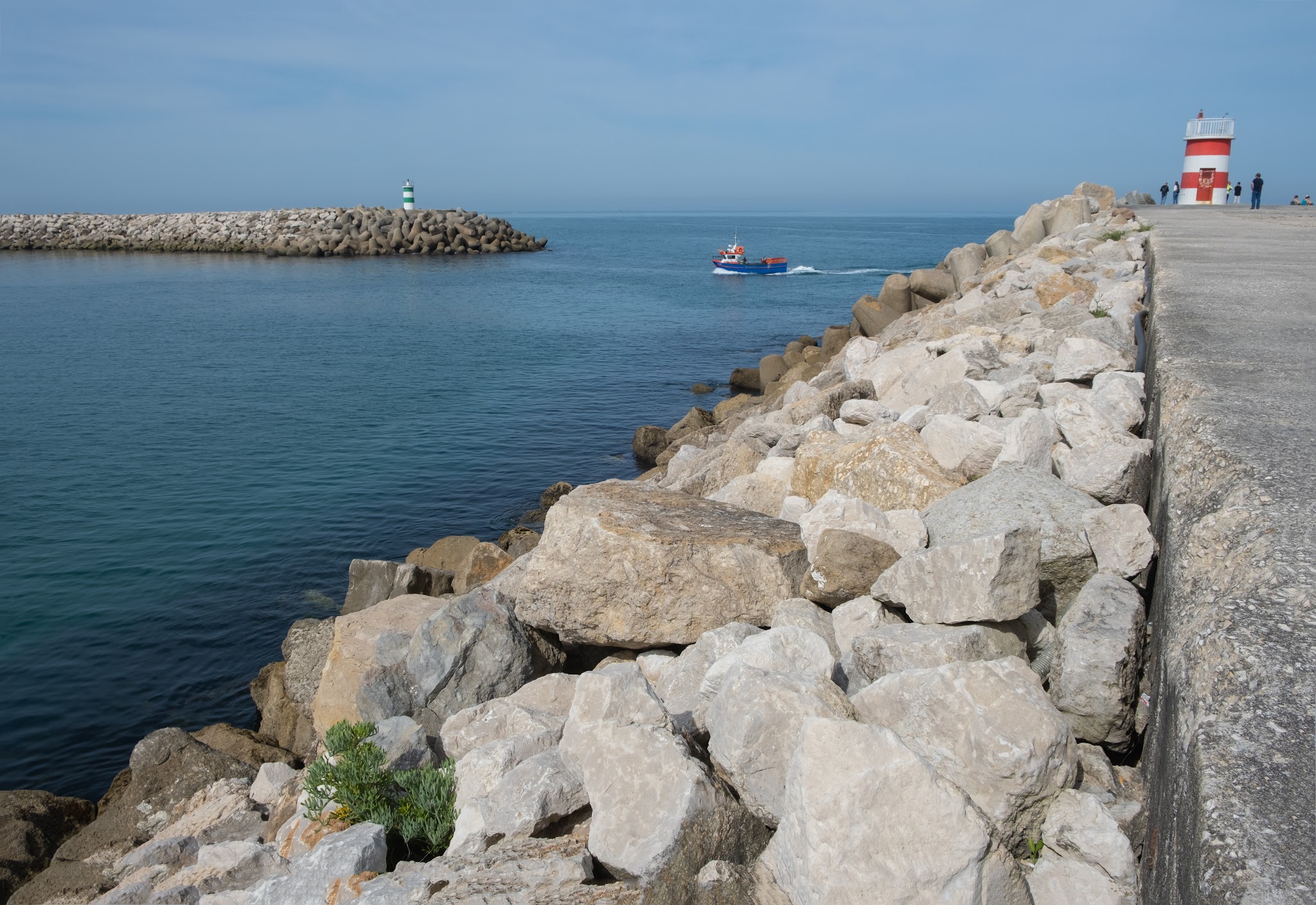 Steinmole mit rot-weißem Leuchtturm rechts; blaues Boot fährt zwischen Mole und Ferndamm mit grünem Leuchtfeuer.