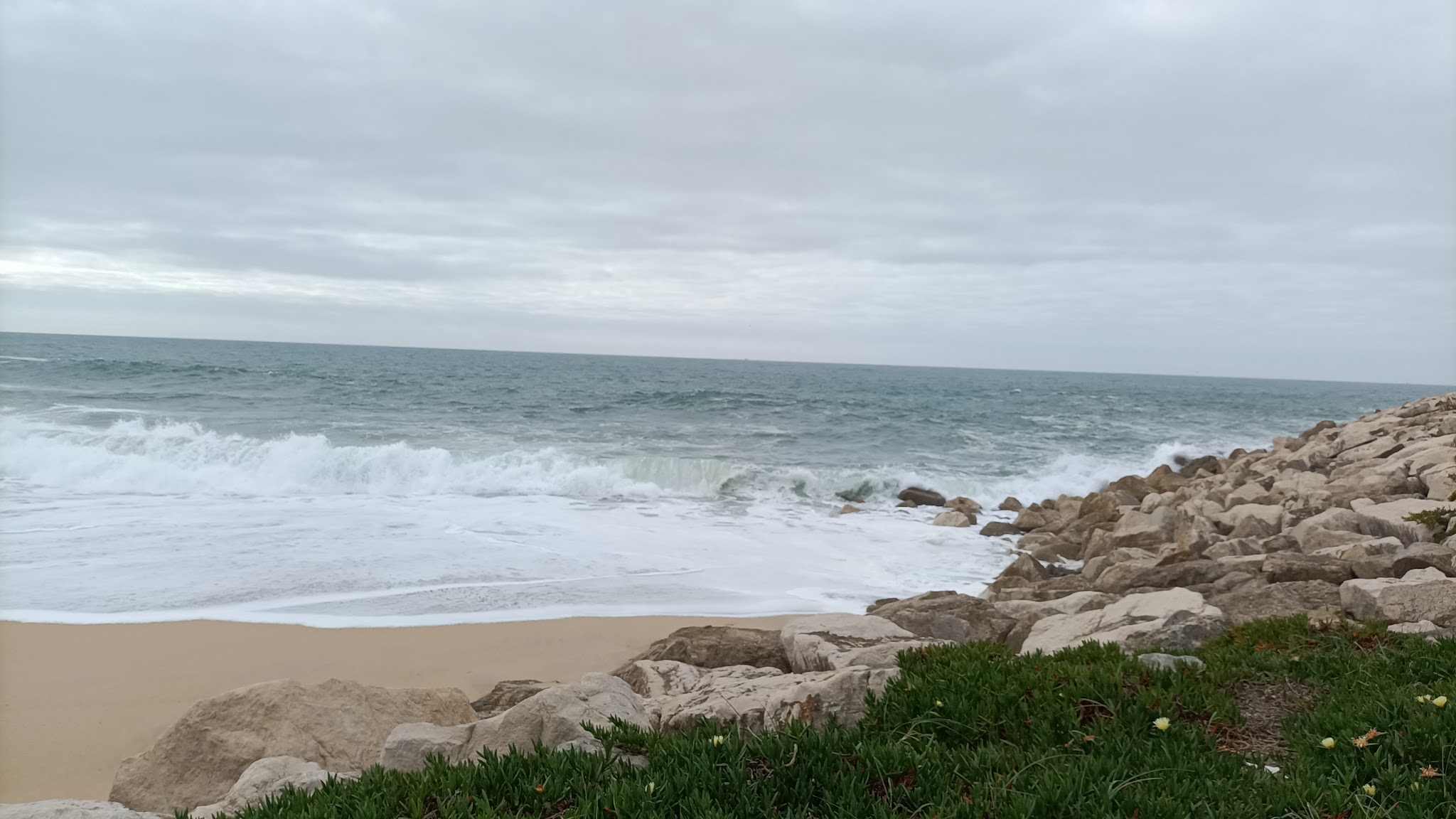 Wellen schlagen an einen sandigen Strand, rechts eine Felsböschung, grauer Himmel.