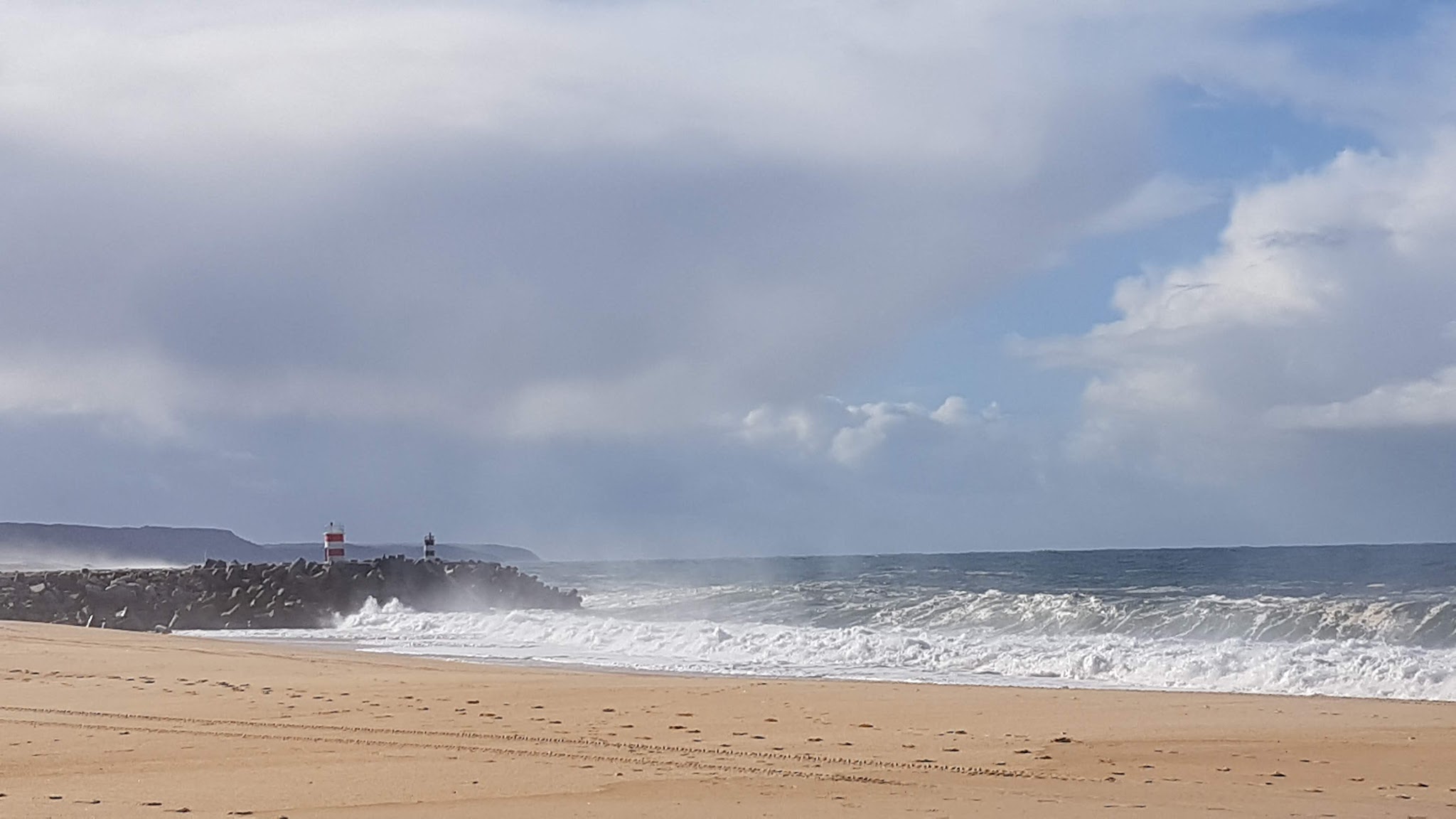 Brandung trifft auf Mole mit rot-weißem Leuchtturm; Sandstrand im Vordergrund unter teils bewölktem Himmel