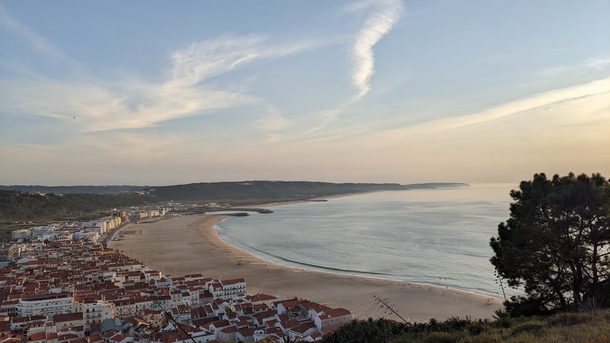 Panoramablick auf ein Küstenstädtchen mit roten Dächern, langer Sandbucht und ruhigem Meer bei sanftem Abendlicht.