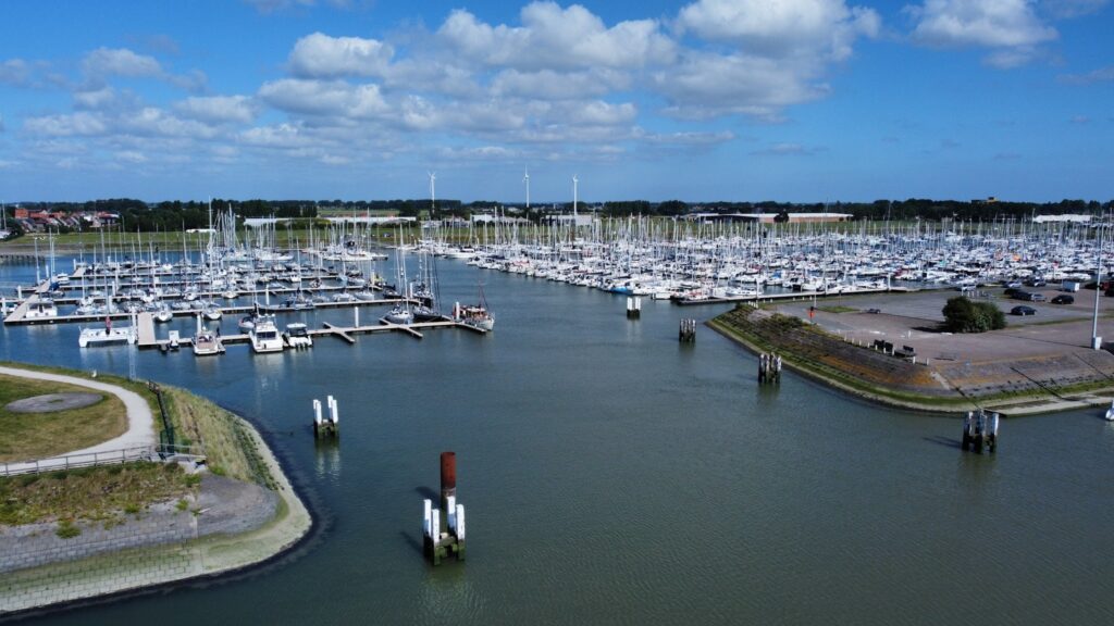 Yachthafen voller Segelboote an ruhigem Wasser; im Hintergrund Windräder und leicht bewölkter Himmel.