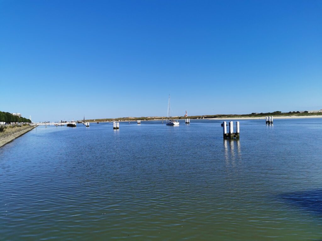Ruhiger Hafenkanal mit vereinzelten Dalben, ankerndem Segelboot und fernem Leuchtturm unter wolkenlosem Himmel.