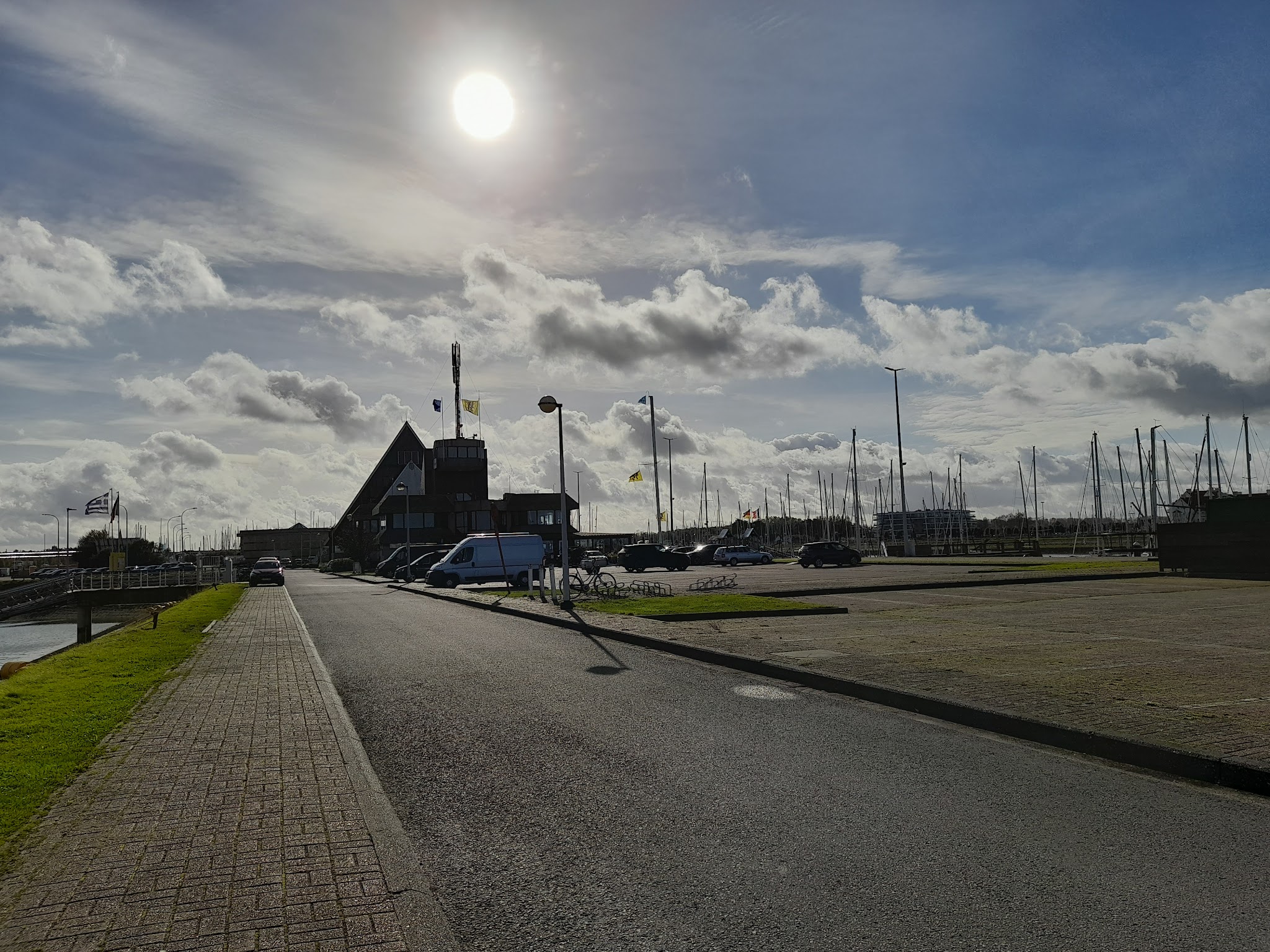 Sonne über Hafen: Straßenblick auf spitzes Hafenhaus, geparkte Autos, Reihe von Segelmasten im Hintergrund.