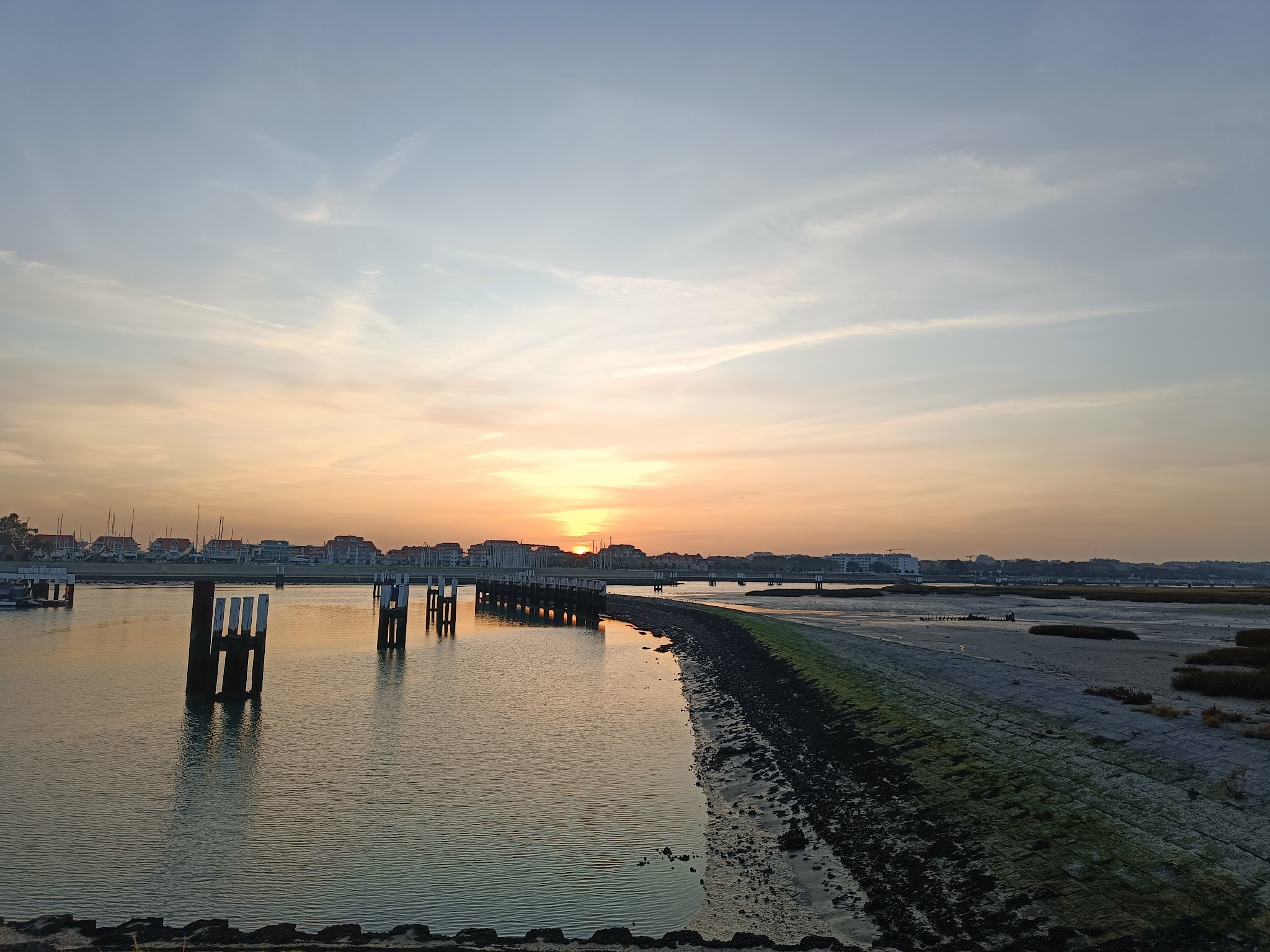 Sonnenuntergang über einem Hafen bei Ebbe, Pfahlreihe und flacher Wasserarm führen zu Häusern am Horizont.