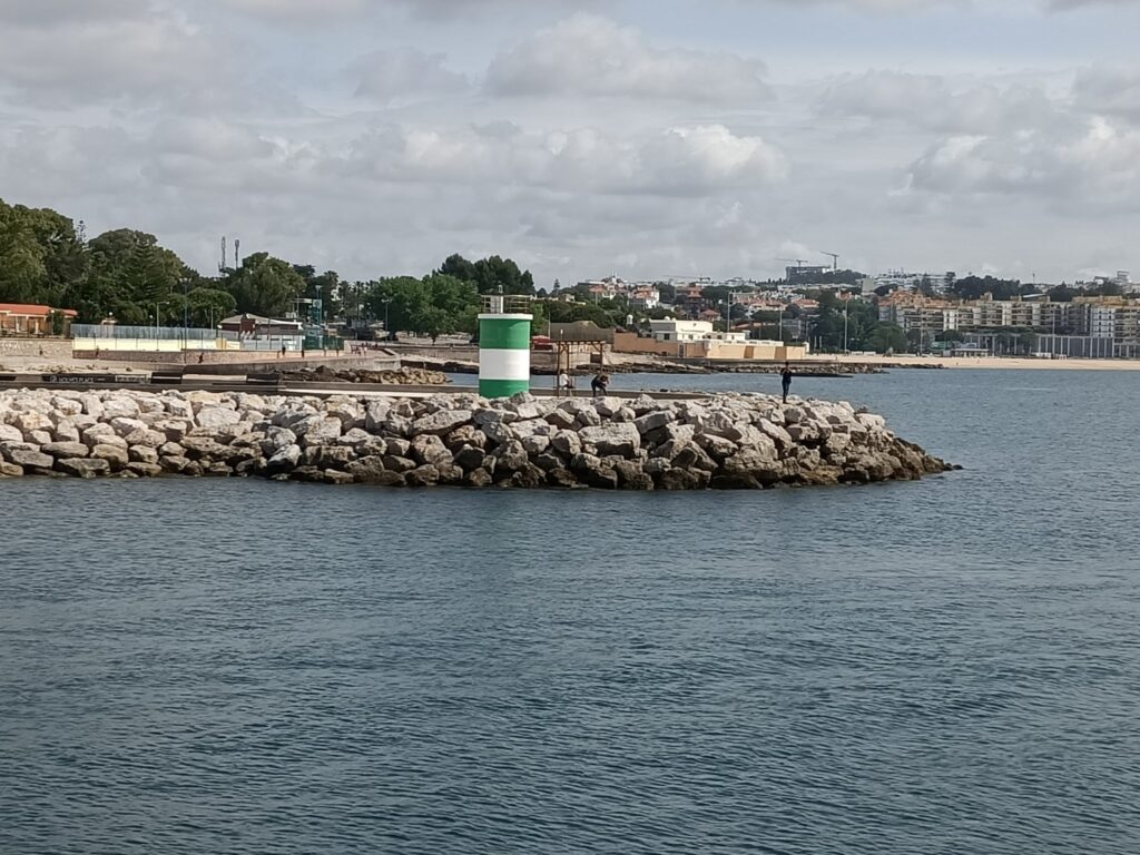 Steinmole mit grün-weißem Leuchtfeuer; zwei Angler blicken aufs ruhige Meer, Stadt und Wolken im Hintergrund