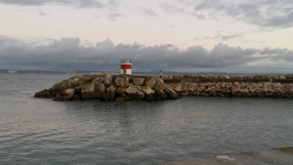 Kleiner rot-weißer Leuchtturm auf Steinmole, Spaziergänger, ruhiges Meer unter bewölktem Abendhimmel.