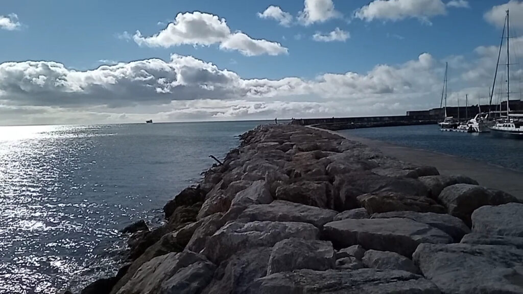 Steinmole führt ins glitzernde Meer; Frachtschiff fern, rechts Segelboote im Hafen, Sonne zwischen Wolken.