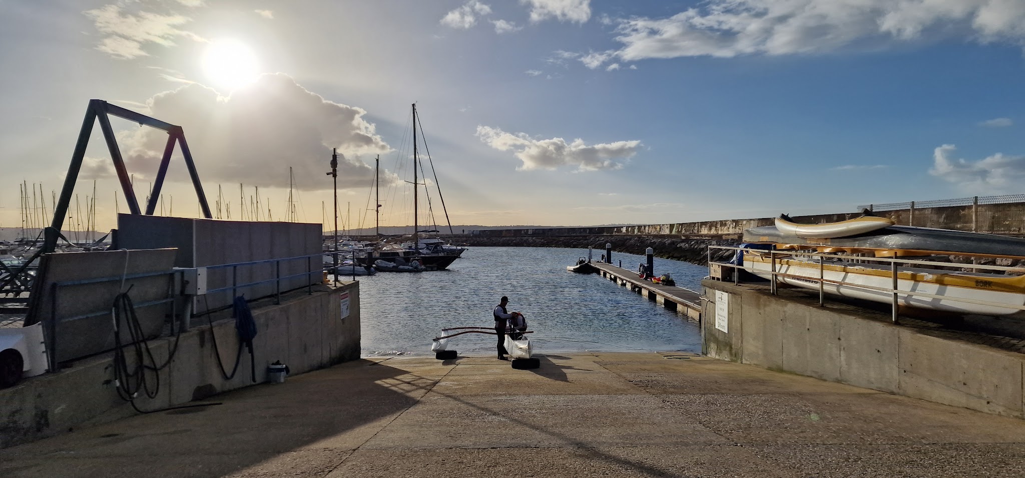 Rampe zum Hafenbecken, Mann richtet kleines Segelboot; Yachten liegen links, Abendsonne blendet durch Wolken.