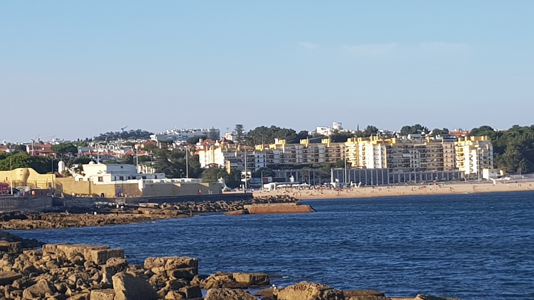 Felsige Küste mit blauem Meer, dahinter Sandstrand und gelb-weiße Apartmenthäuser unter klarem Himmel