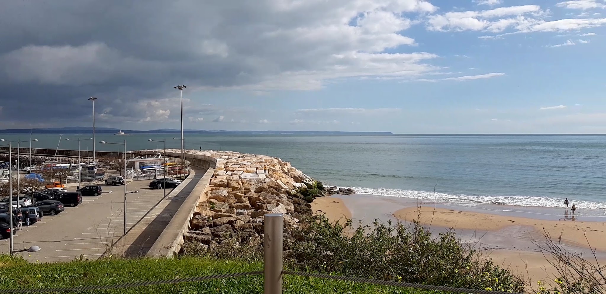 Felsmole mit Parkplatz, daneben sandiger Strand; zwei Spaziergänger im seichten Meer, wechselhaft bewölkter Himmel