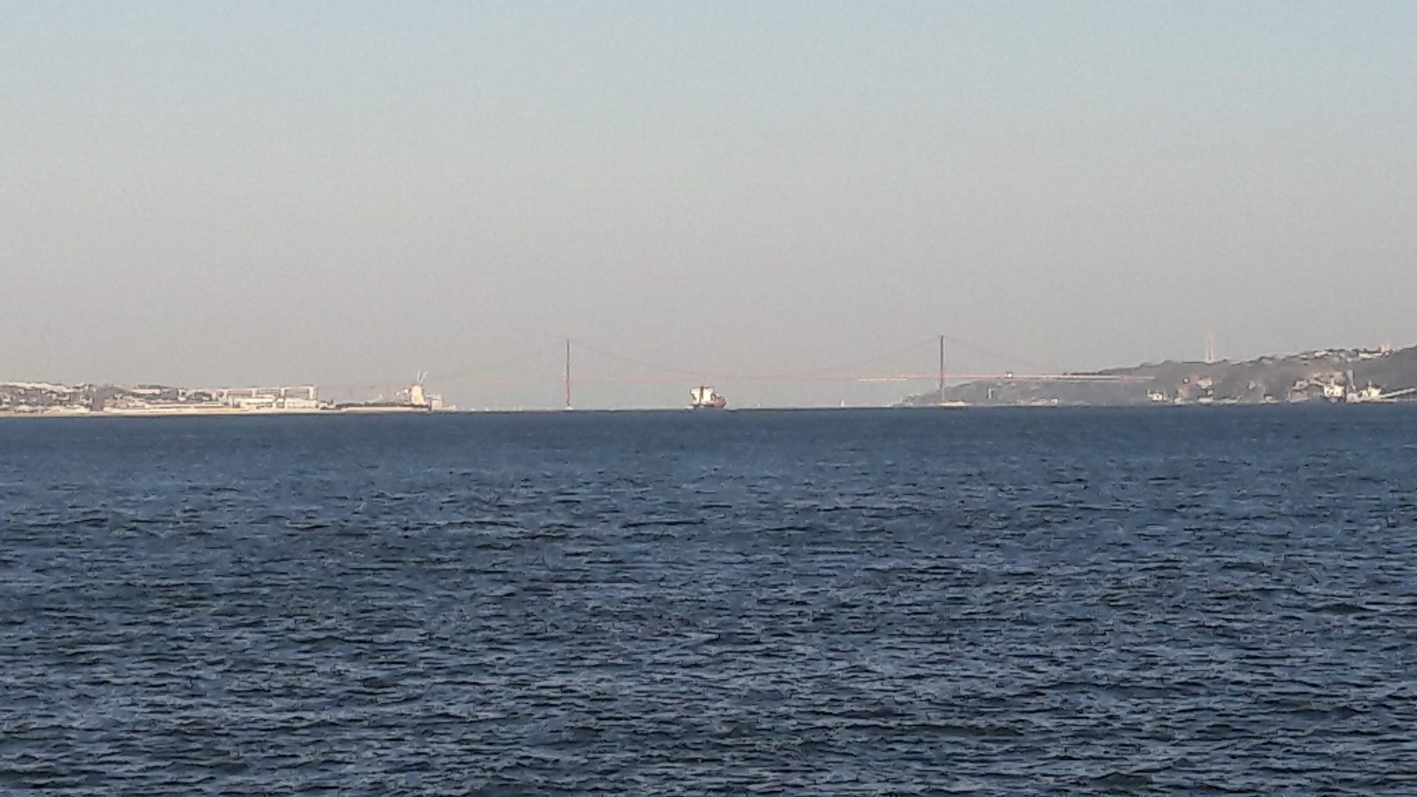 Weites blaues Wasser, ferne Küste mit roter Hängebrücke; Frachtschiff fährt unter der Brücke.