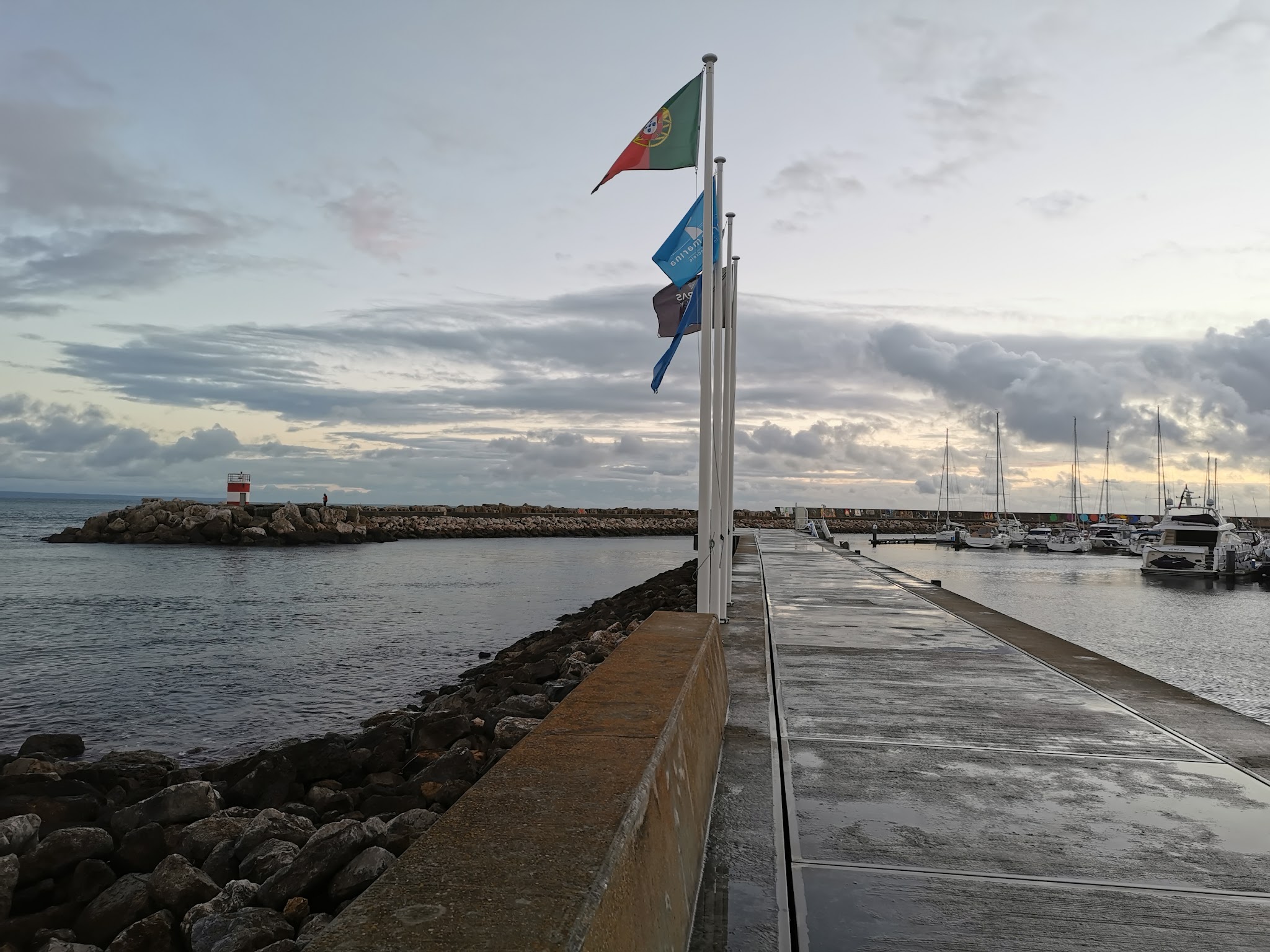 Nasser Steg mit wehenden Flaggen führt zu Yachthafen; links Wellenbrecher mit kleinem Leuchtturm unter wolkigem Himmel.
