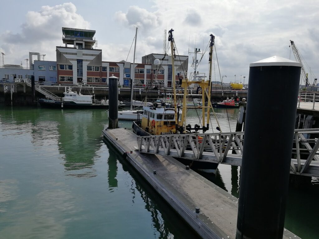 Gelbes Fischereiboot am schwimmenden Steg im Hafen; dahinter moderne Lagerhäuser, Masten und Hafenkräne.