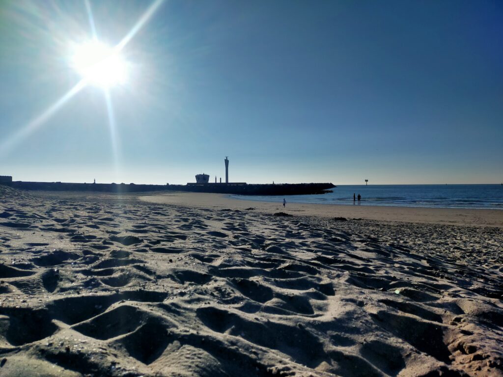 Strand bei tiefstehender Sonne, strukturierter Sand im Vordergrund, wenige Menschen und Mole mit Leuchtturm am Horizont.