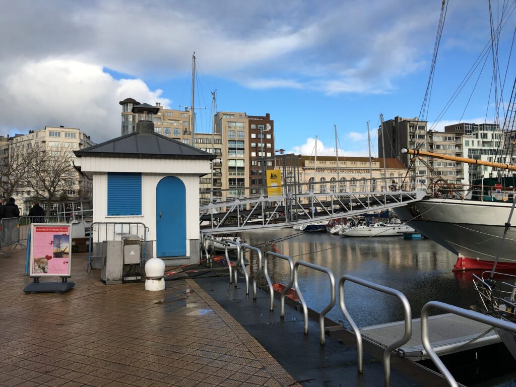 Kleiner weiß-blauer Hafenpavillon neben Gangway zu Segelschiff; moderne Häuser und Boote spiegeln sich im Wasser.