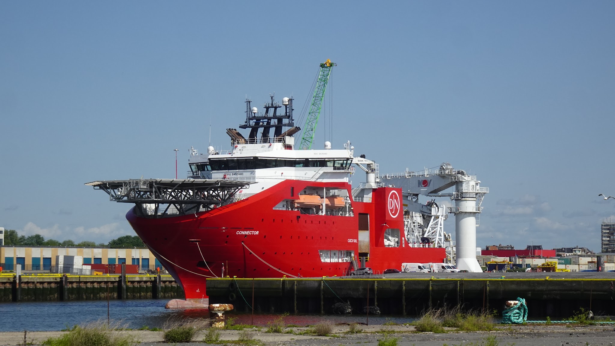 Großes rotes Versorgungsschiff mit Helikopterdeck liegt am Kai, Kran und Hafenanlagen im Hintergrund.