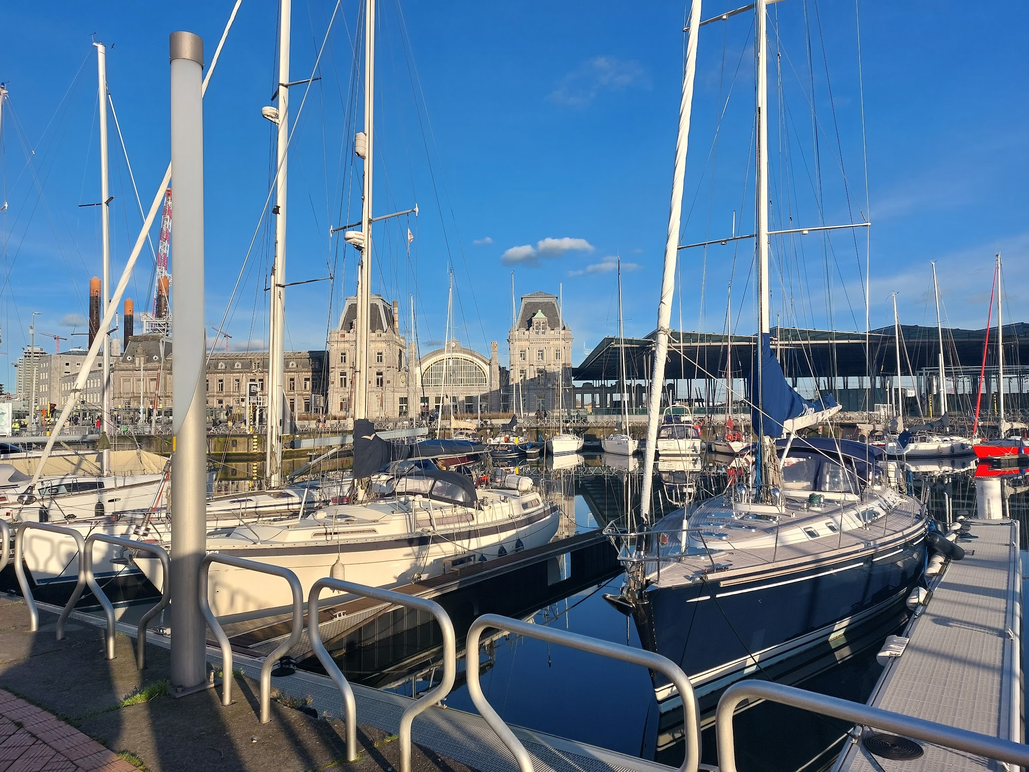 Segelyachten im sonnigen Hafen vor Amsterdams Centraal-Bahnhof, Masten spiegeln sich im ruhigen Wasser.