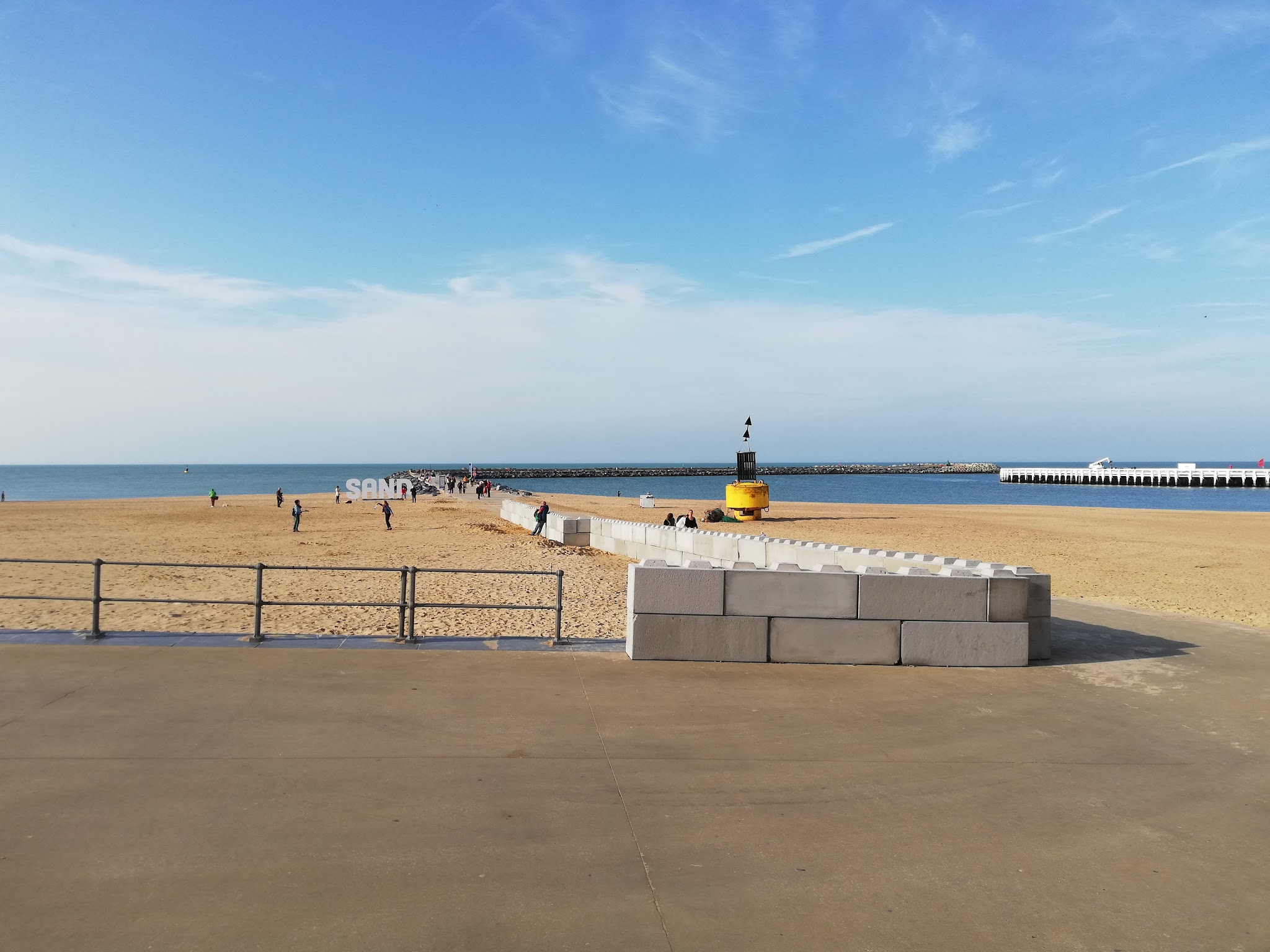 Weitläufiger Strand mit vereinzelten Spaziergängern, Steinblockweg zur Mole, Schriftzug ‚SAND‘, ruhiges Meer und blauer Himmel