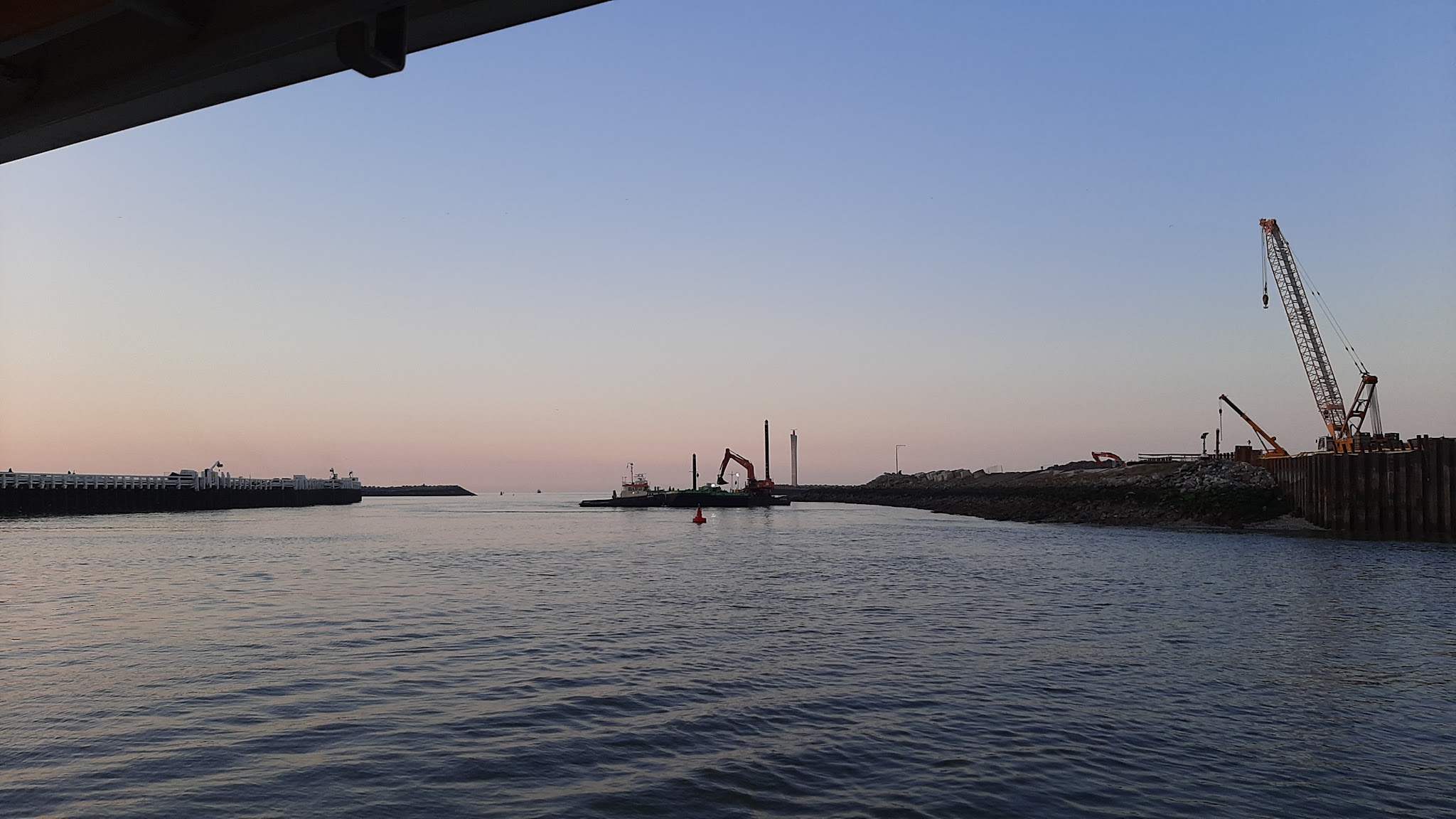 Hafeneinfahrt im Abendlicht, ruhiges Wasser, Baggerponton und zwei Kräne arbeiten an beiden Ufern.