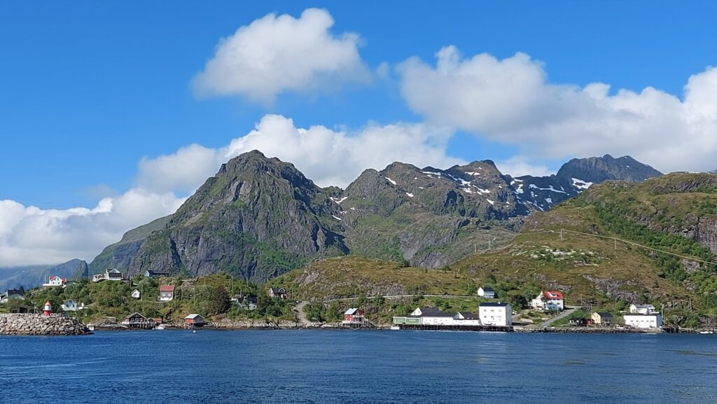 Norwegisches Küstendorf mit bunten Häusern und kleinem Leuchtturm vor hohen, grünen Bergen mit Schneeflecken