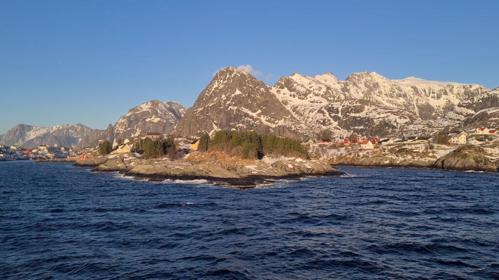 Schneebedeckte Berge hinter bunten Holzhäusern auf felsiger Küste; vorn dunkelblaues, bewegtes Meer unter klarem Himmel.