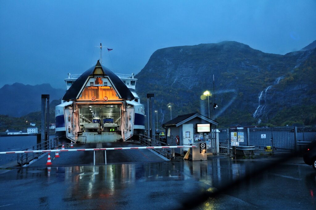 Abendlich beleuchtete Autofähre mit geöffneter Bugklappe am regennassen Kai, Berge und Wasserfälle im Hintergrund.