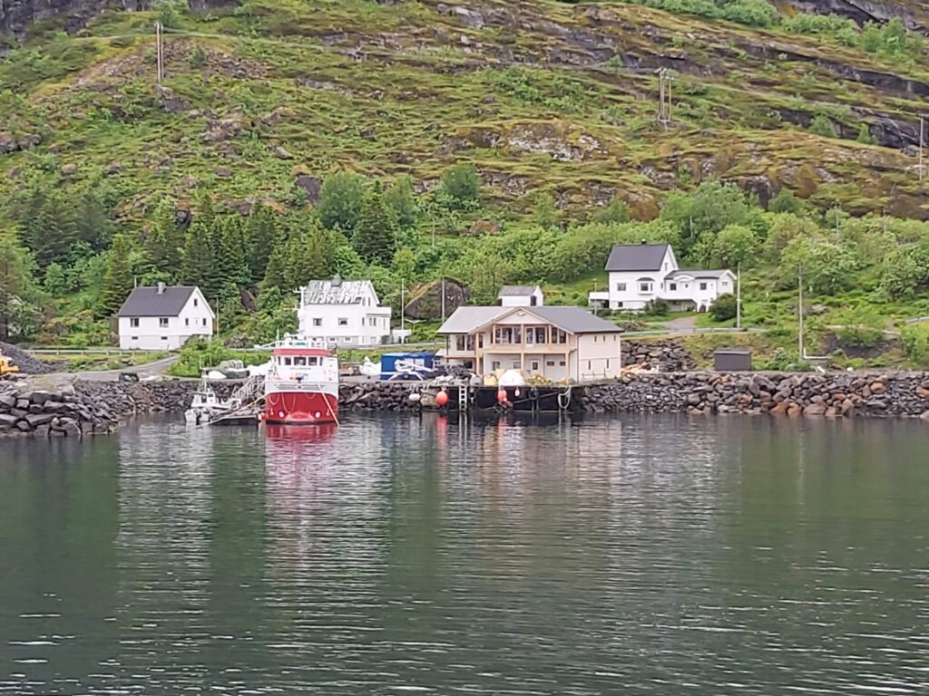 Kleiner Fjordhafen: rotes Ausflugsschiff an Steg, umgeben von weißen Holzhäusern und grünen Berghängen