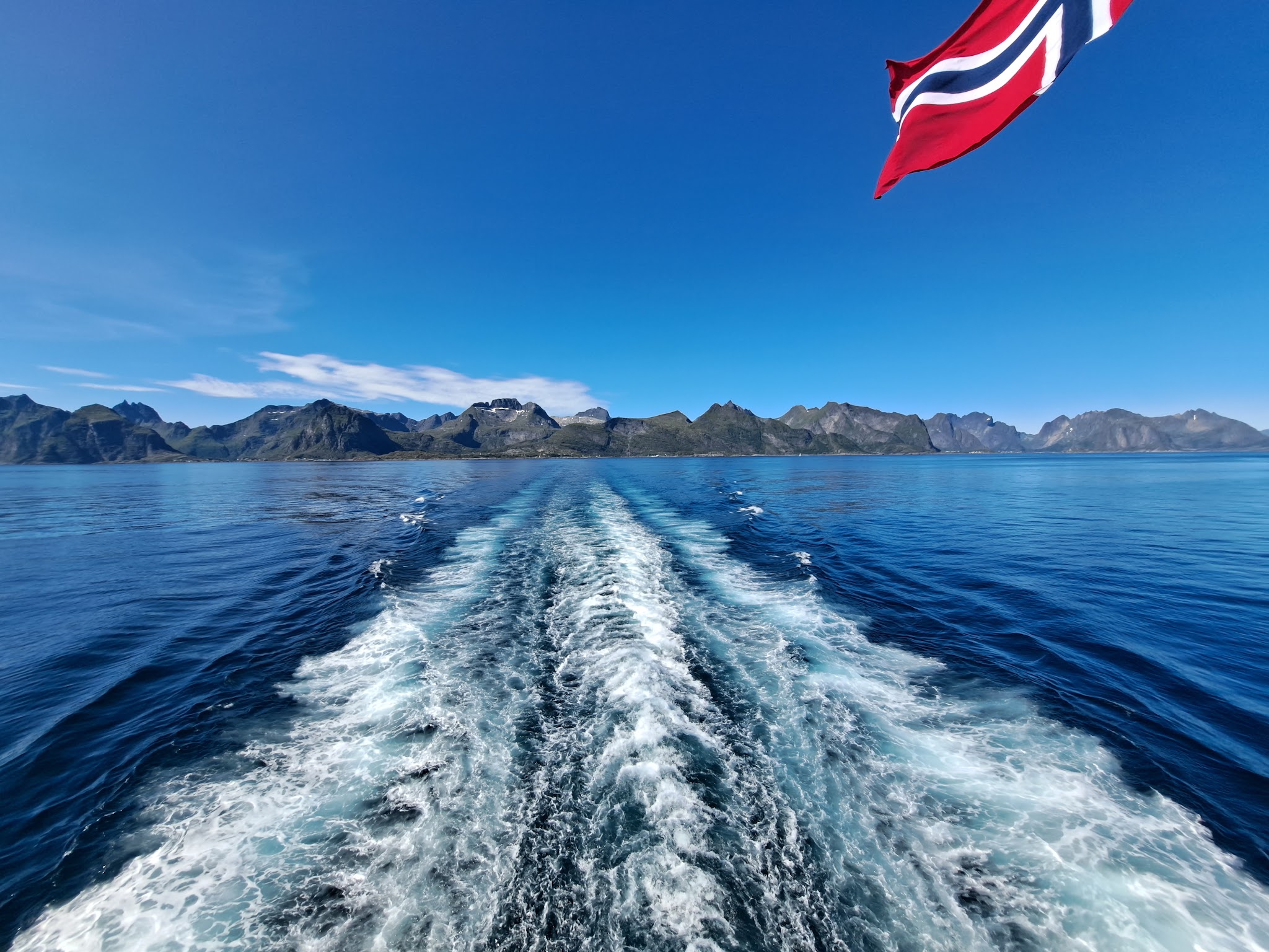 Bootsgischt zieht durchs tiefblaue Meer, Norwegische Flagge weht oben rechts, fern zerklüftete Küstenberge.