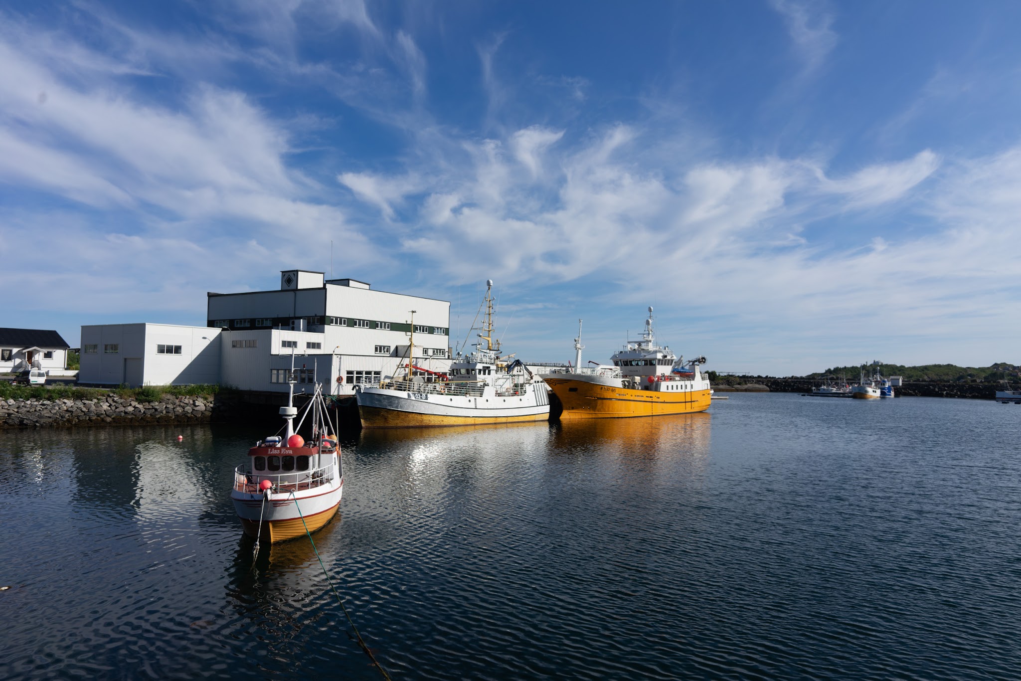Drei Fischerboote, zwei gelb und eins rot-weiß, vertäut vor weißem Hafengebäude unter klarem blauen Himmel