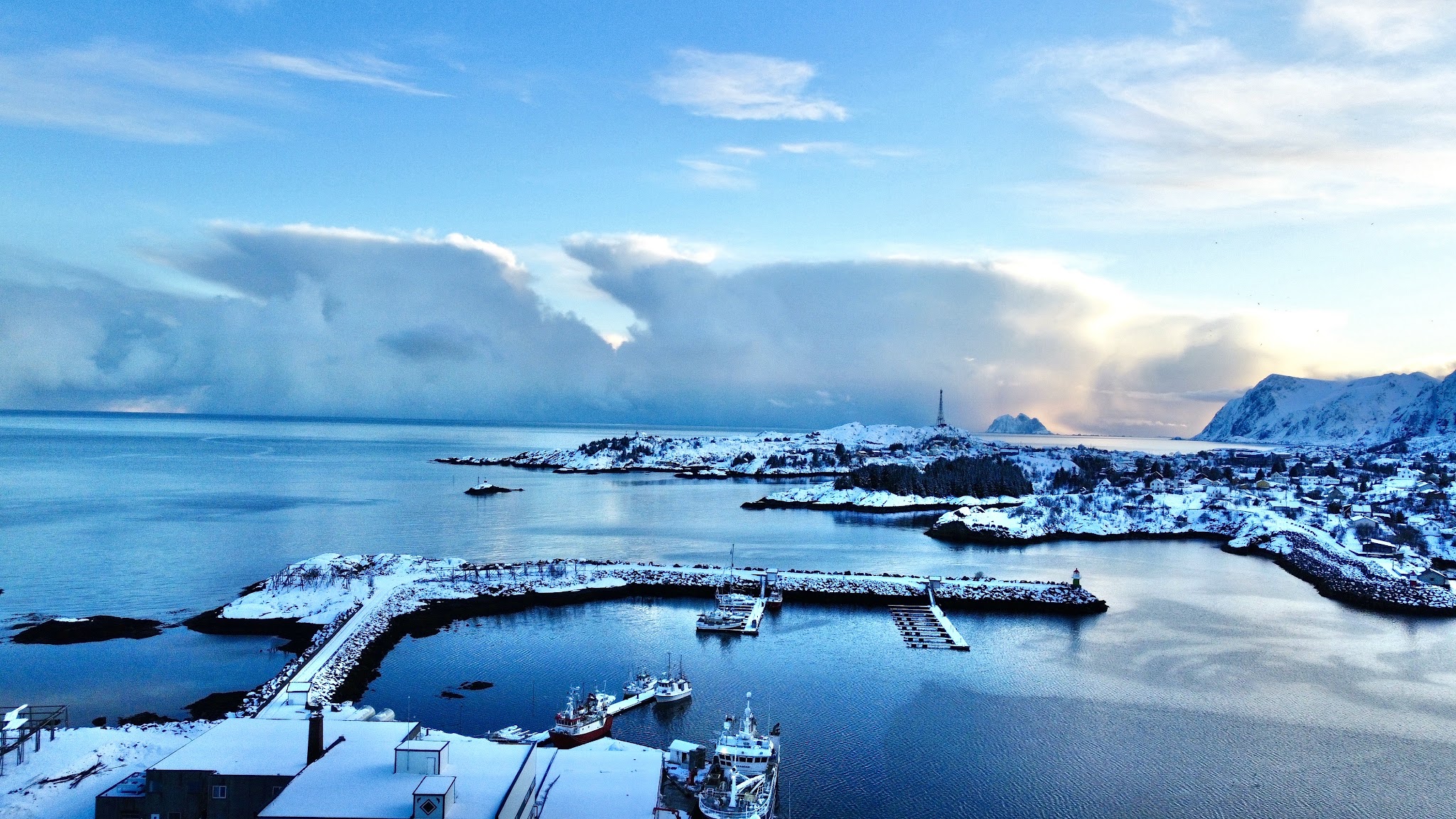 Verschneiter Küstenort mit kleinem Hafen und Fischerbooten; ruhiges Meer unter dramatischem Morgenhimmel.