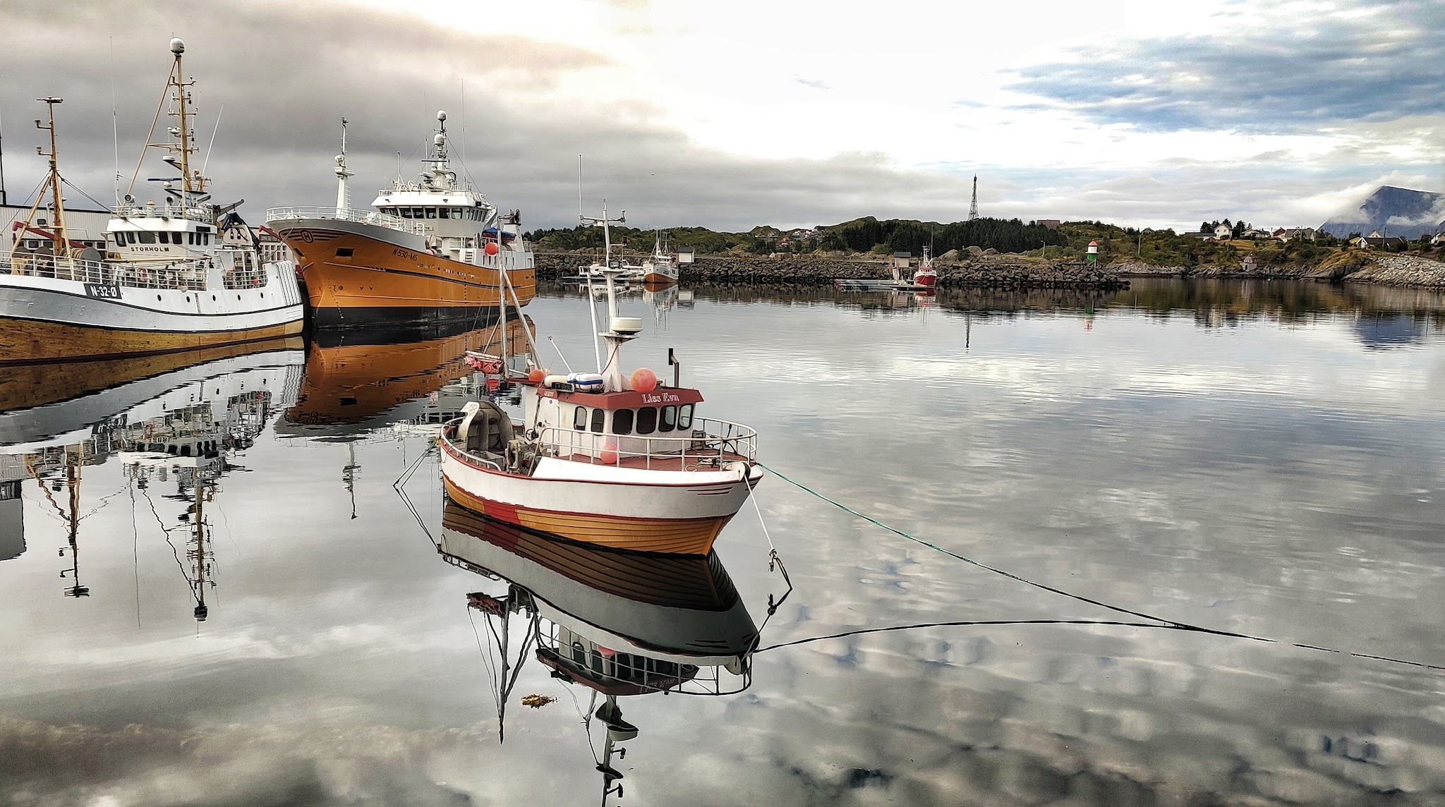 Leicht bewölkter Hafen: kleines buntes Fischerboot spiegelt sich im ruhigen Wasser, zwei größere Kutter dahinter.