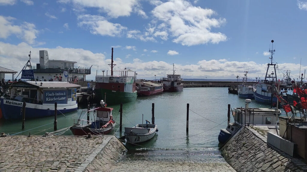 Kleiner Hafen mit mehreren Fischerbooten am Kai, ruhiges Wasser, darüber blauer Himmel mit Schäfchenwolken.